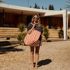 Femme debout pieds nus devant une maison moderne ensoleillée, portant un grand sac cabas rayé orange et blanc, des lunettes de soleil blanches et un sweat foncé.