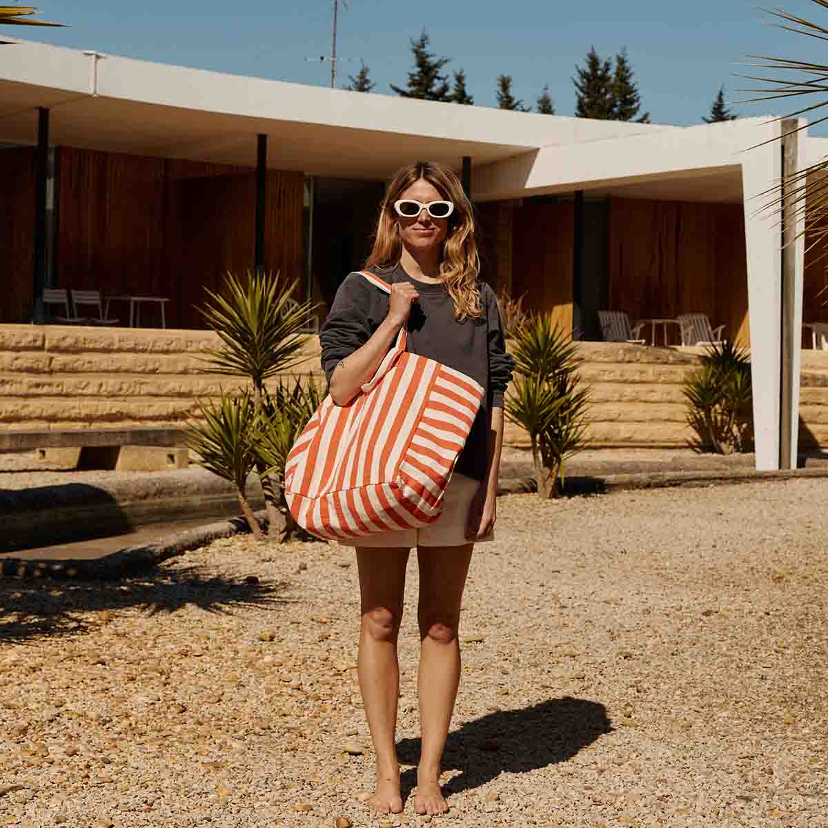 Femme debout pieds nus devant une maison moderne ensoleillée, portant un grand sac cabas rayé orange et blanc, des lunettes de soleil blanches et un sweat foncé.