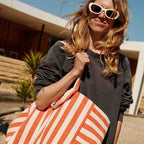 Gros plan d’une femme portant des lunettes de soleil blanches et un sweat foncé, tenant un grand sac cabas rayé orange et blanc devant une maison moderne ensoleillée.