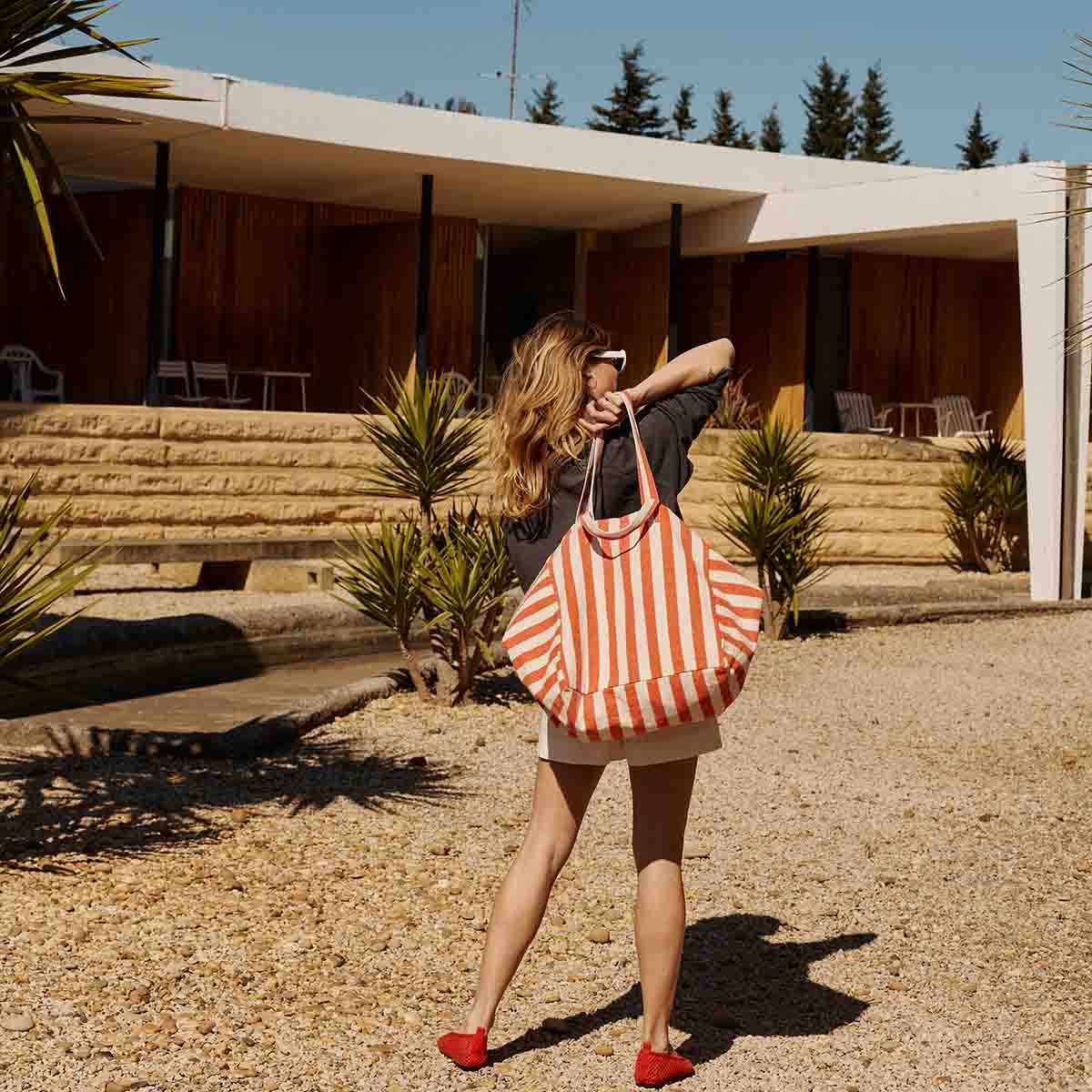 Femme de dos devant une maison moderne ensoleillée. Elle porte le sac à une main dans son dos. 

