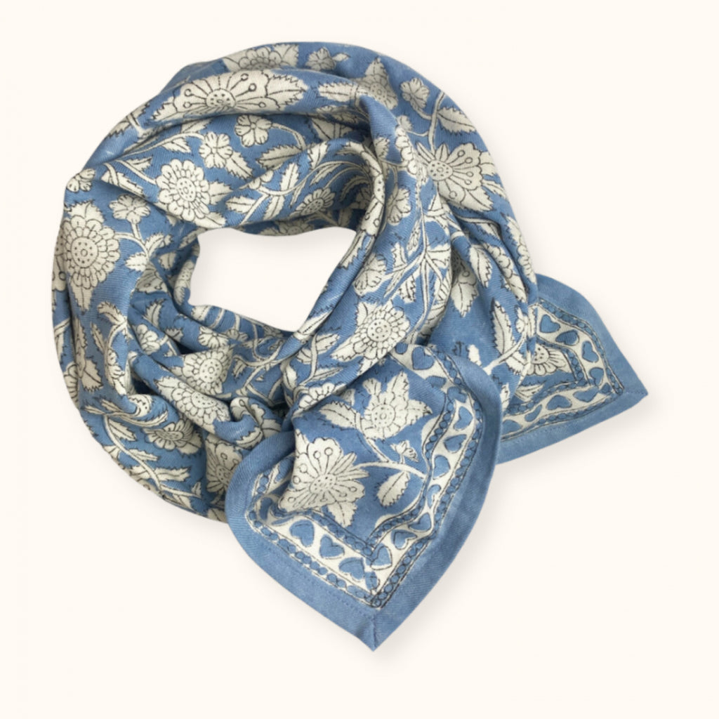 Le foulard est noué et à motifs. On y voit des fleures blanches au contour noir fin  sur un fond bleu ciel. La bordure du foulard à un autre motif représenté par des coeur bleu ciel sur un fond bleu ciel. 