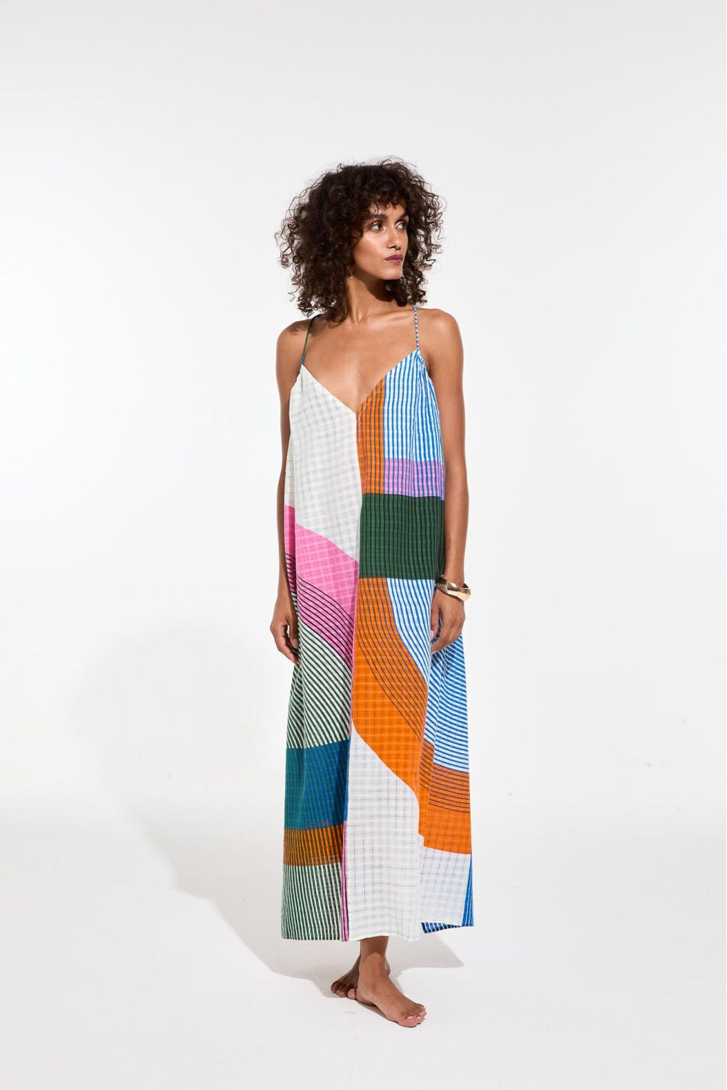 Une femme porte une longue robe géométrique. la robe est dans les tons bleu ciel, vert, rose, blanc et orange avec des lignes ou des carreaux. 