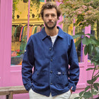 un homme brun porte une veste de couleur marine style marin. Il se tient droit avec les mains dans les poches. La veste est droite, elle est composée d' un col chemise, de manches longues avec une coupe droite. il l'a porte fermée grâce aux 5 boutons placés à la vertical mais il a laissé les deux premiers ouverts. Il y a également des écussons, l'un se trouve sur l'une des poches plaquées au niveau de ses hanches et l'autre sur l'extrémité de la manche. 