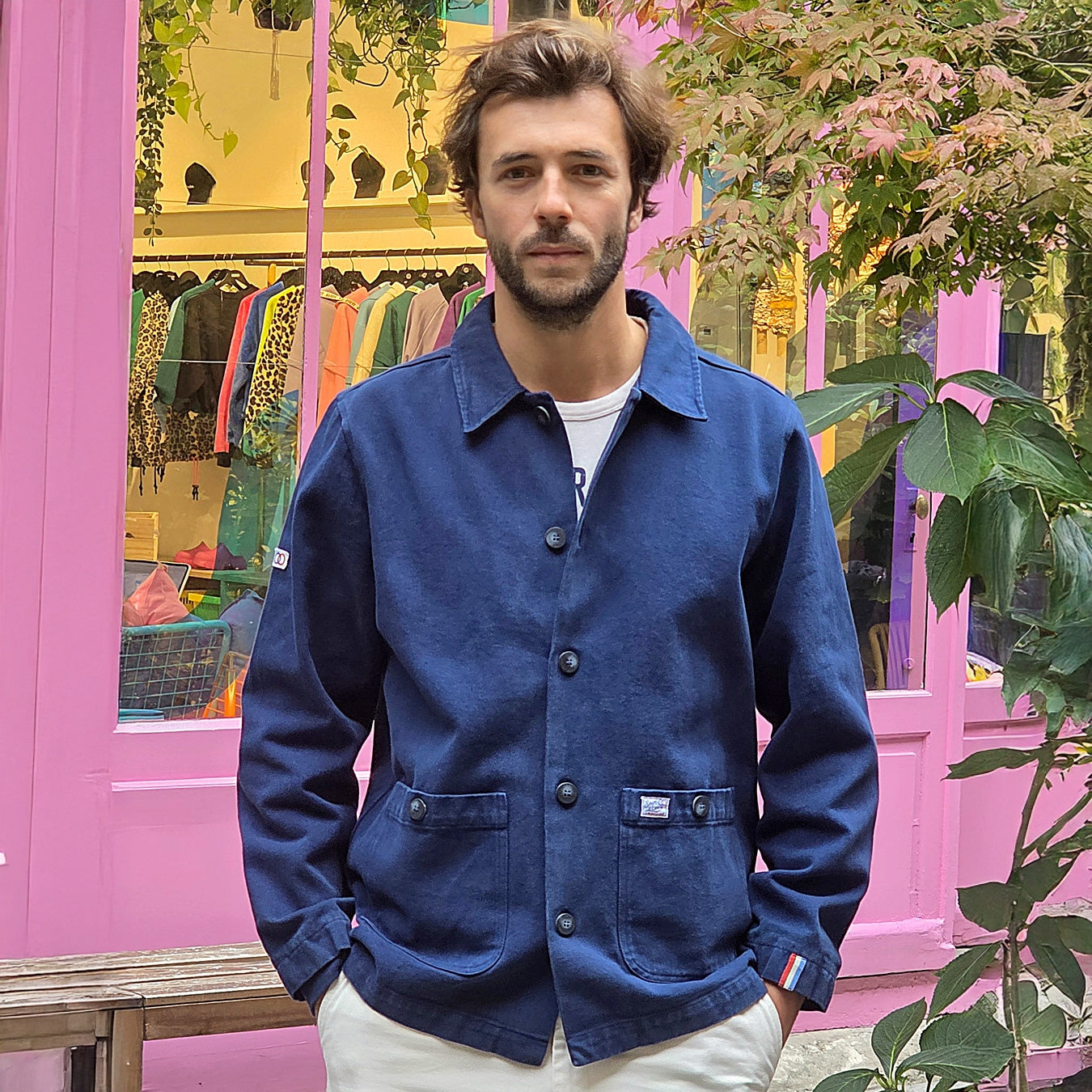 un homme brun porte une veste de couleur marine style marin. Il se tient droit avec les mains dans les poches. La veste est droite, elle est composée d' un col chemise, de manches longues avec une coupe droite. il l'a porte fermée grâce aux 5 boutons placés à la vertical mais il a laissé les deux premiers ouverts. Il y a également des écussons, l'un se trouve sur l'une des poches plaquées au niveau de ses hanches et l'autre sur l'extrémité de la manche. 